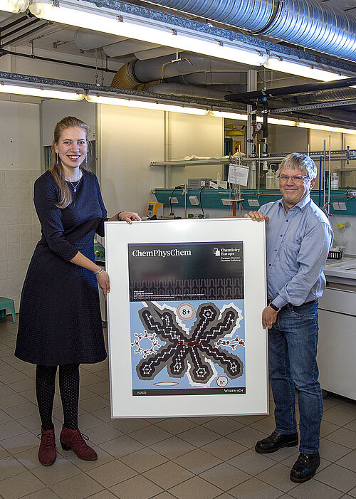 Masterstudentin Jule Philipp und Professor Ralf Ludwig mit der aktuellen Titelseite der Fachzeitschrift für Physikalische Chemie. Das Motiv zeigt ein Oktagon aus Molekül-Ionen. (Foto: Universität Rostock/Julia Tetzke).