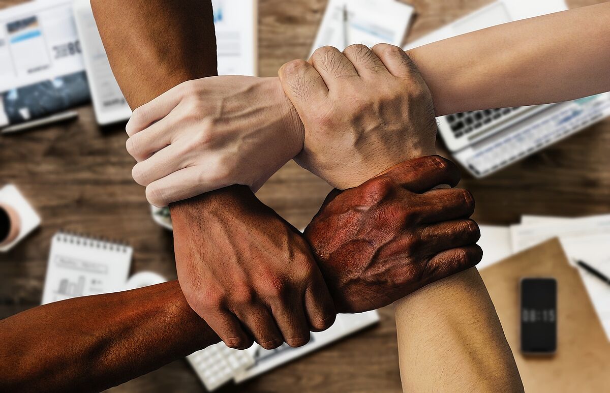 Four arms of different skin color reach out from the sides of the picture to the center. The hands each grab the wrist of the next hand, creating a square.